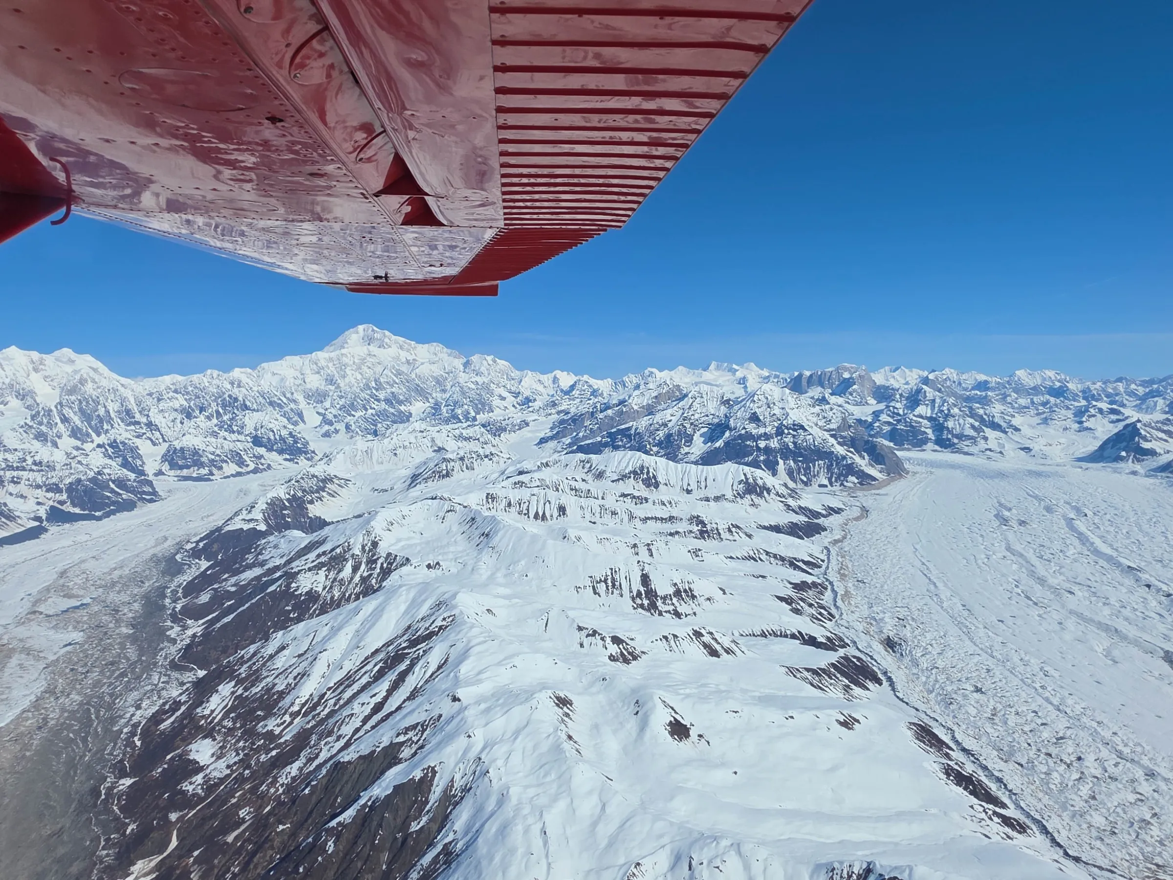 Aerial view of snowy mountains and glaciers under a wing of a red airplane.