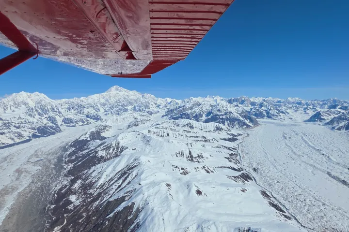 Aerial view of snowy mountains and glaciers under a wing of a red airplane.