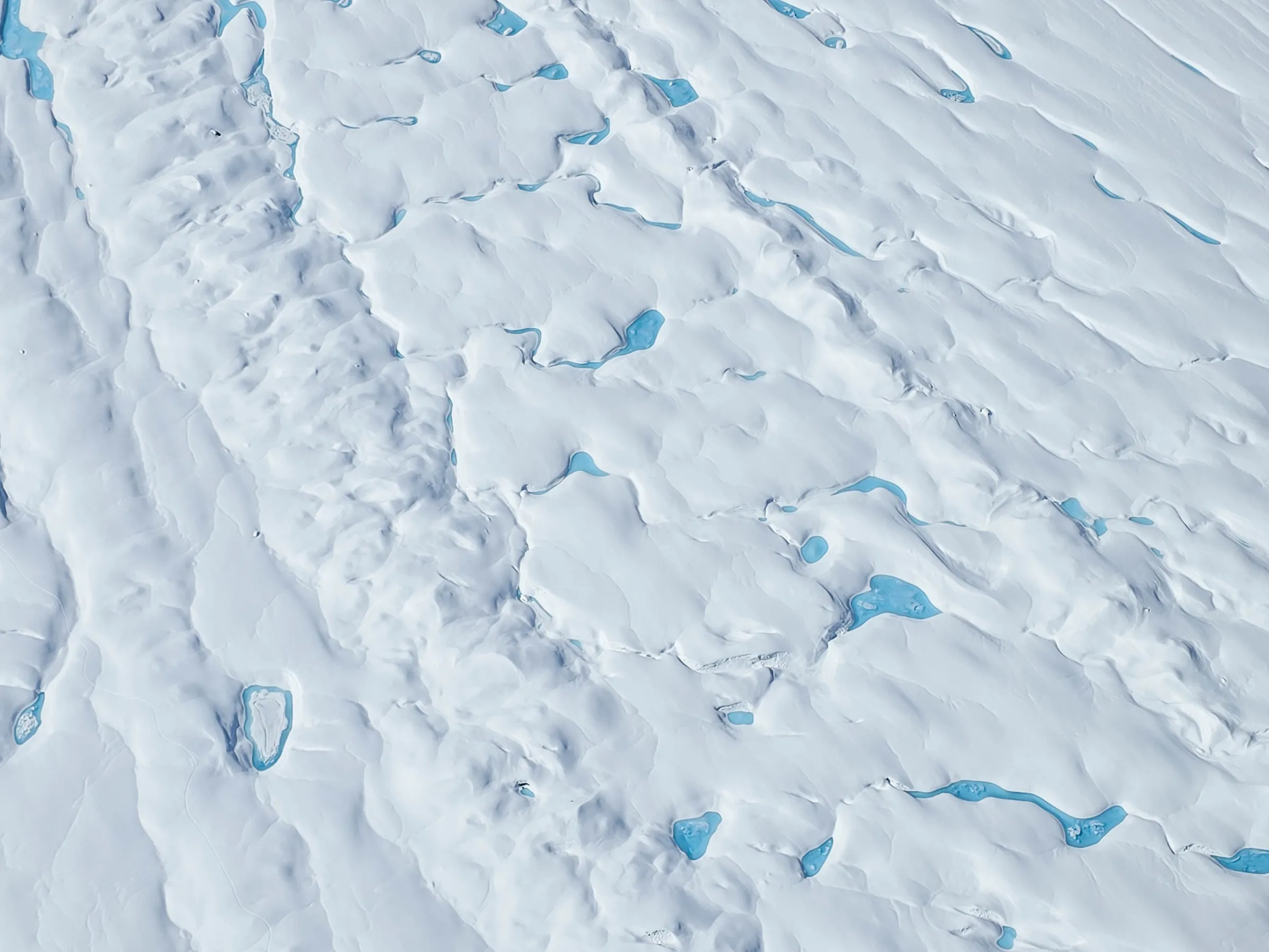 Aerial view of snowy landscape with scattered blue melt ponds.