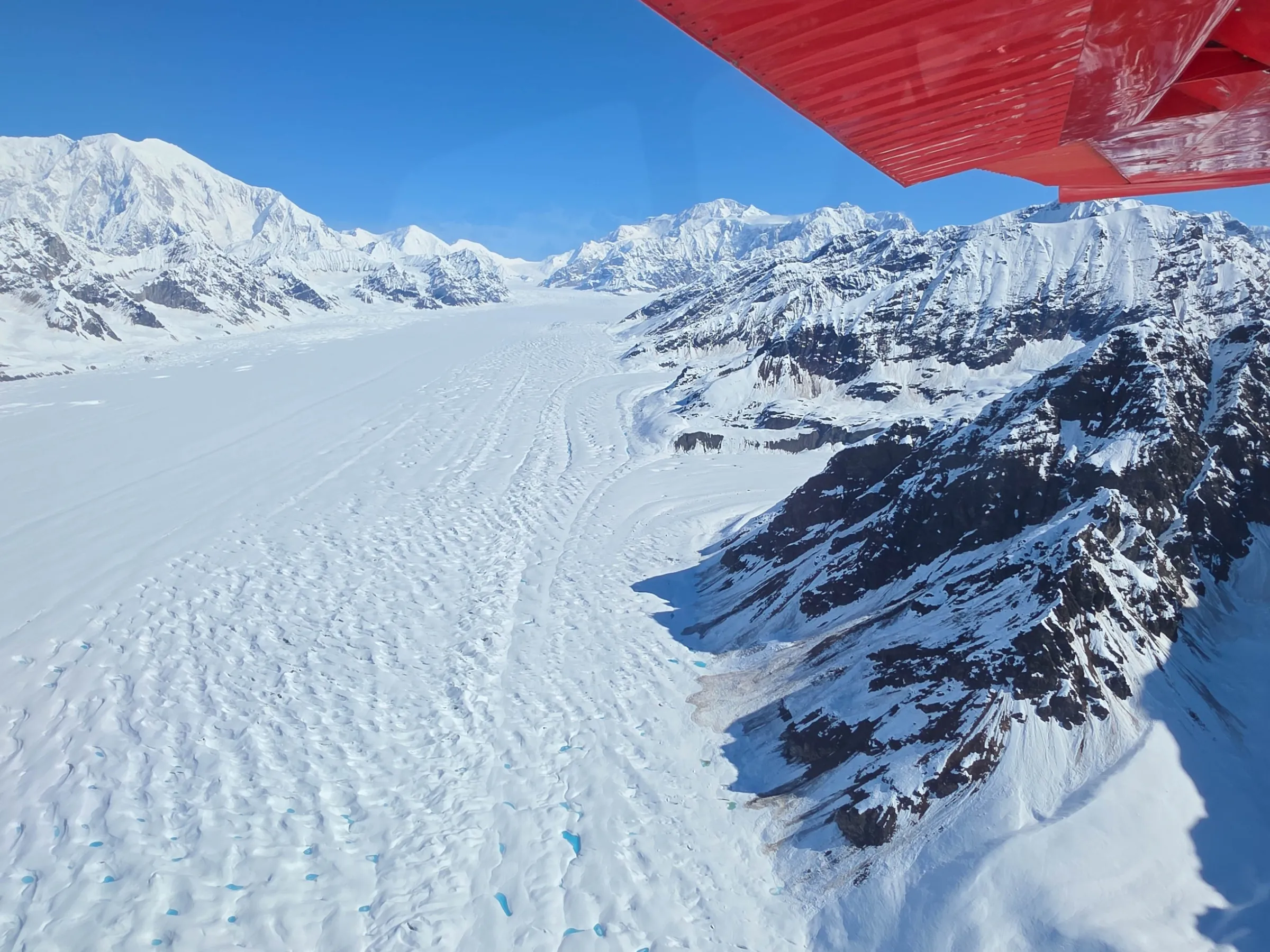 Aerial view of a vast snowy glacier with mountains, under a blue sky and a red plane wing visible.