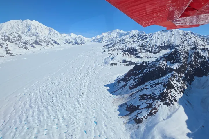 Aerial view of a vast snowy glacier with mountains, under a blue sky and a red plane wing visible.