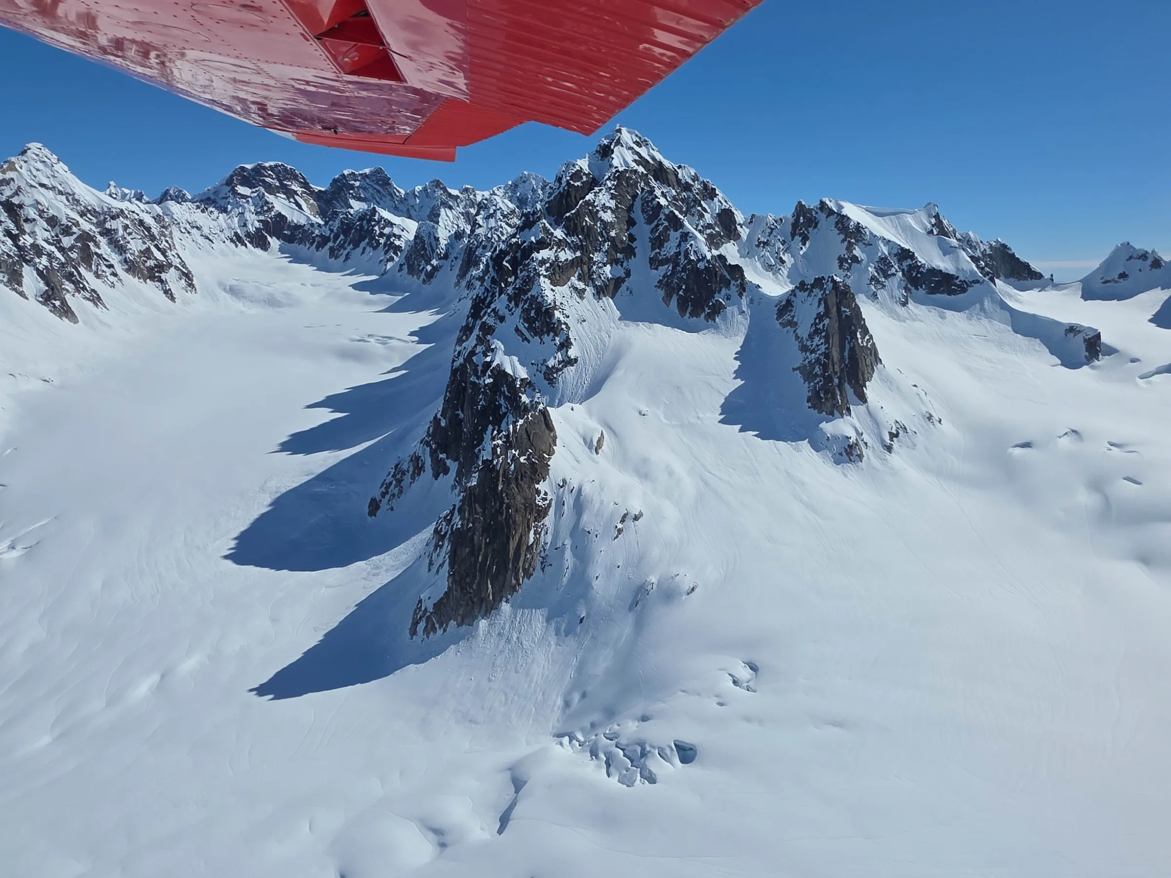 Snow-covered mountain peaks under a clear blue sky with a red airplane wing in view.