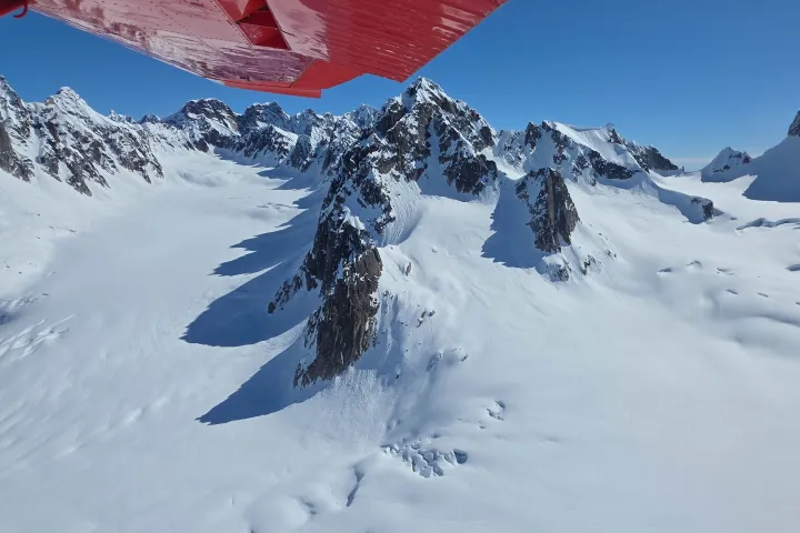 Snow-covered mountain peaks under a clear blue sky with a red airplane wing in view.