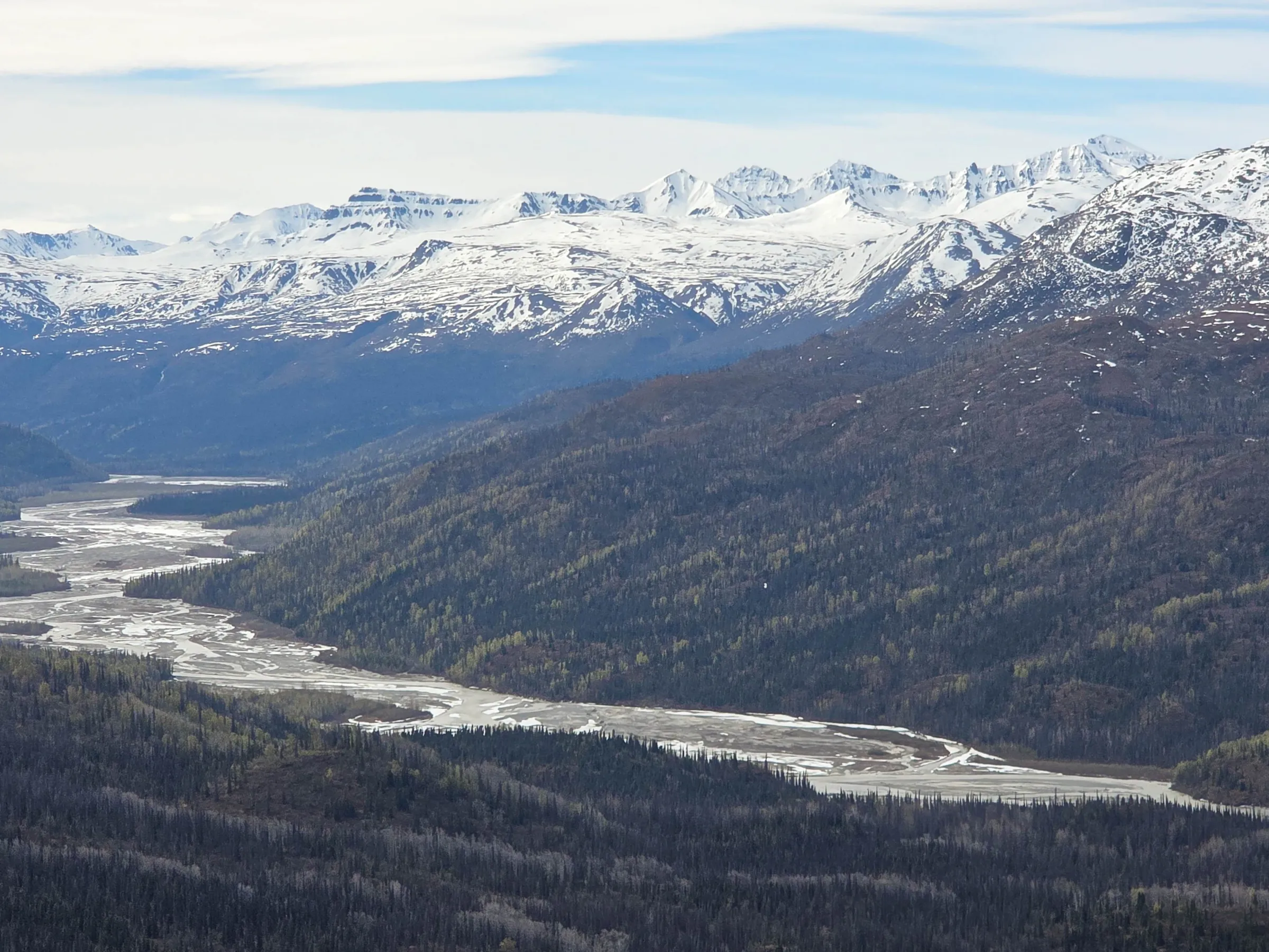 Mountain range with snow peaks and river winding through forested valley.