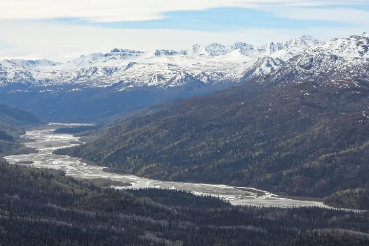 Mountain range with snow peaks and river winding through forested valley.