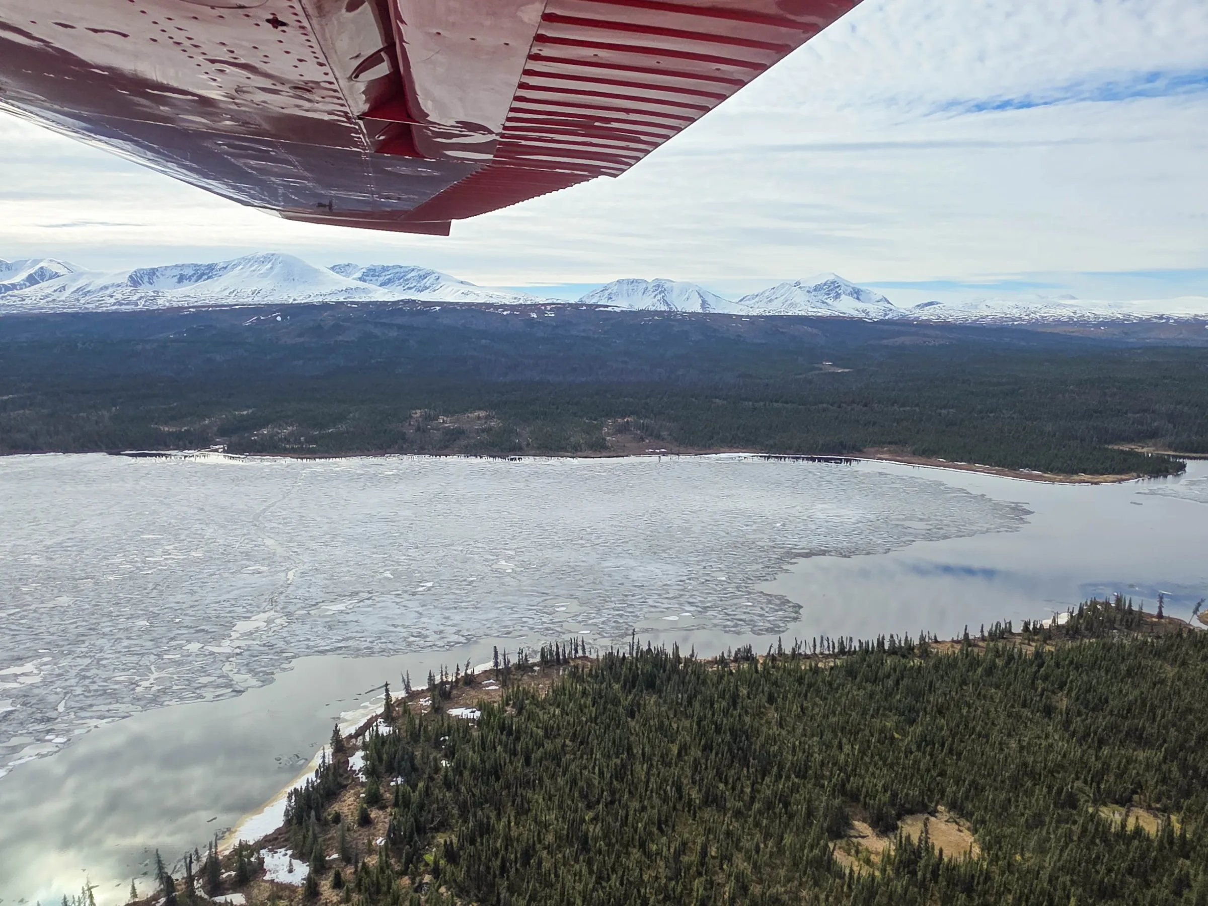 View of snowy mountains, frozen lake, and forest from a plane's wing.