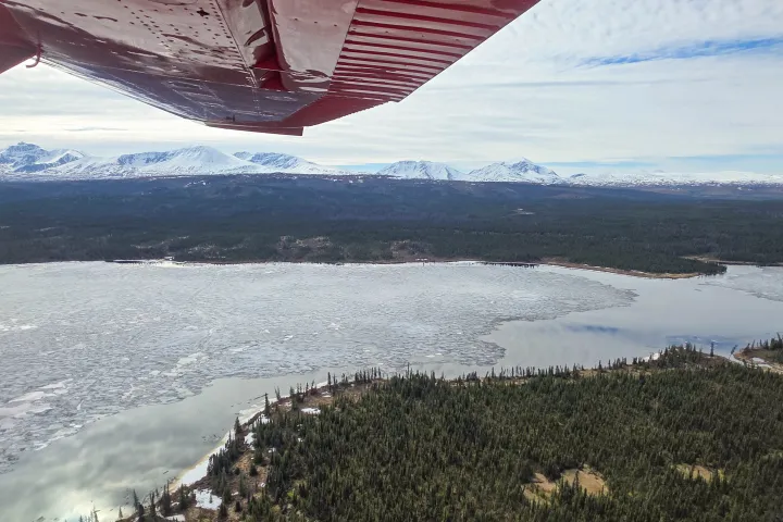 View of snowy mountains, frozen lake, and forest from a plane's wing.