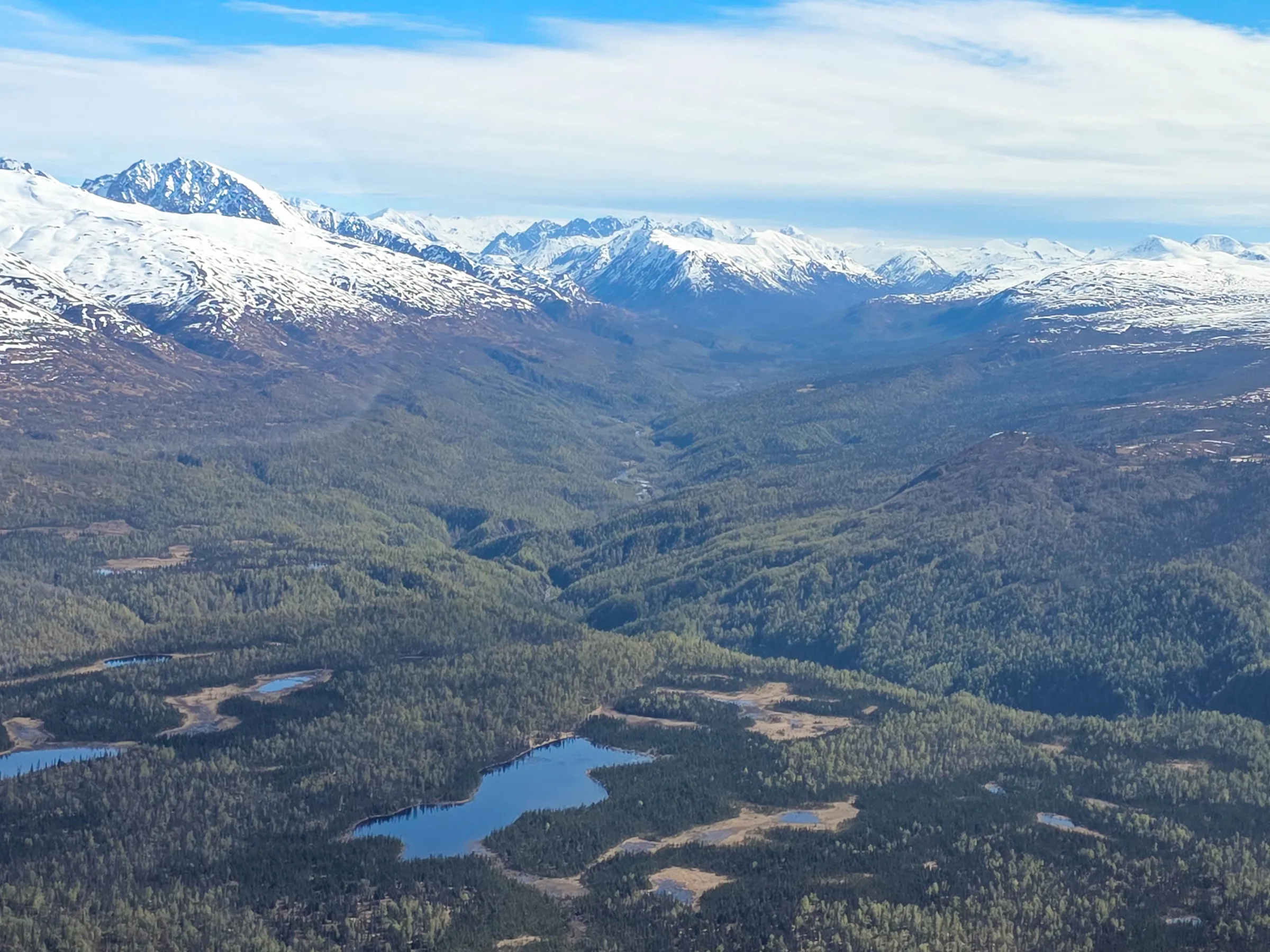 Aerial view of snow-capped mountains, a green valley, and small lakes under a cloudy sky.