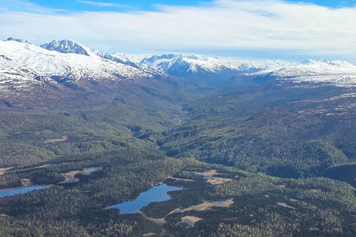 Aerial view of snow-capped mountains, a green valley, and small lakes under a cloudy sky.