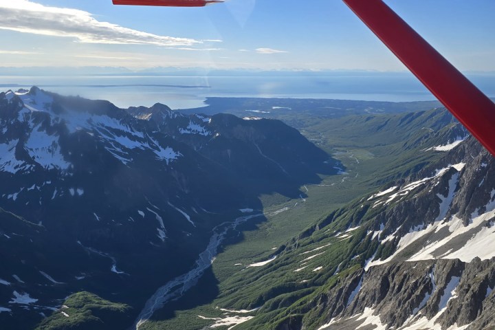 Aerial view of a mountainous valley with snow patches, under a red plane wing.