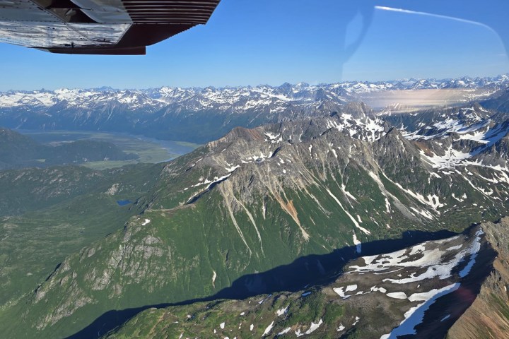 Aerial view of snow-capped mountains and green valleys under a clear blue sky, with part of an airplane wing visible.