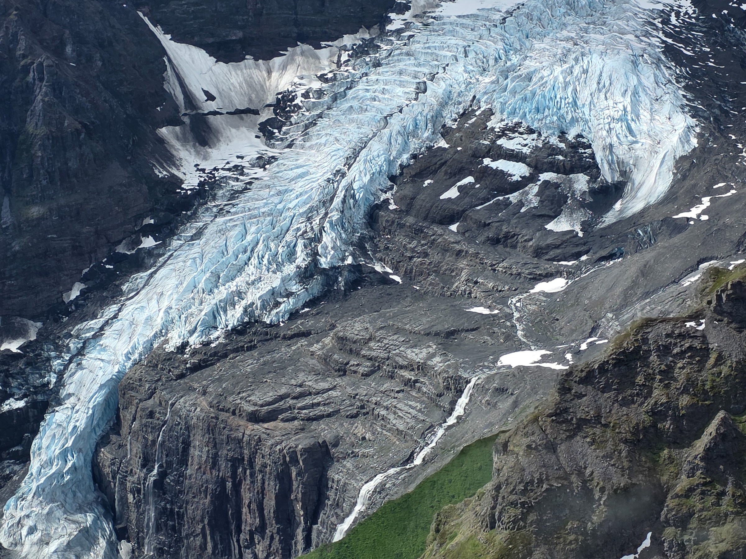 A glacier flowing between rocky mountains with patches of snow and greenery.