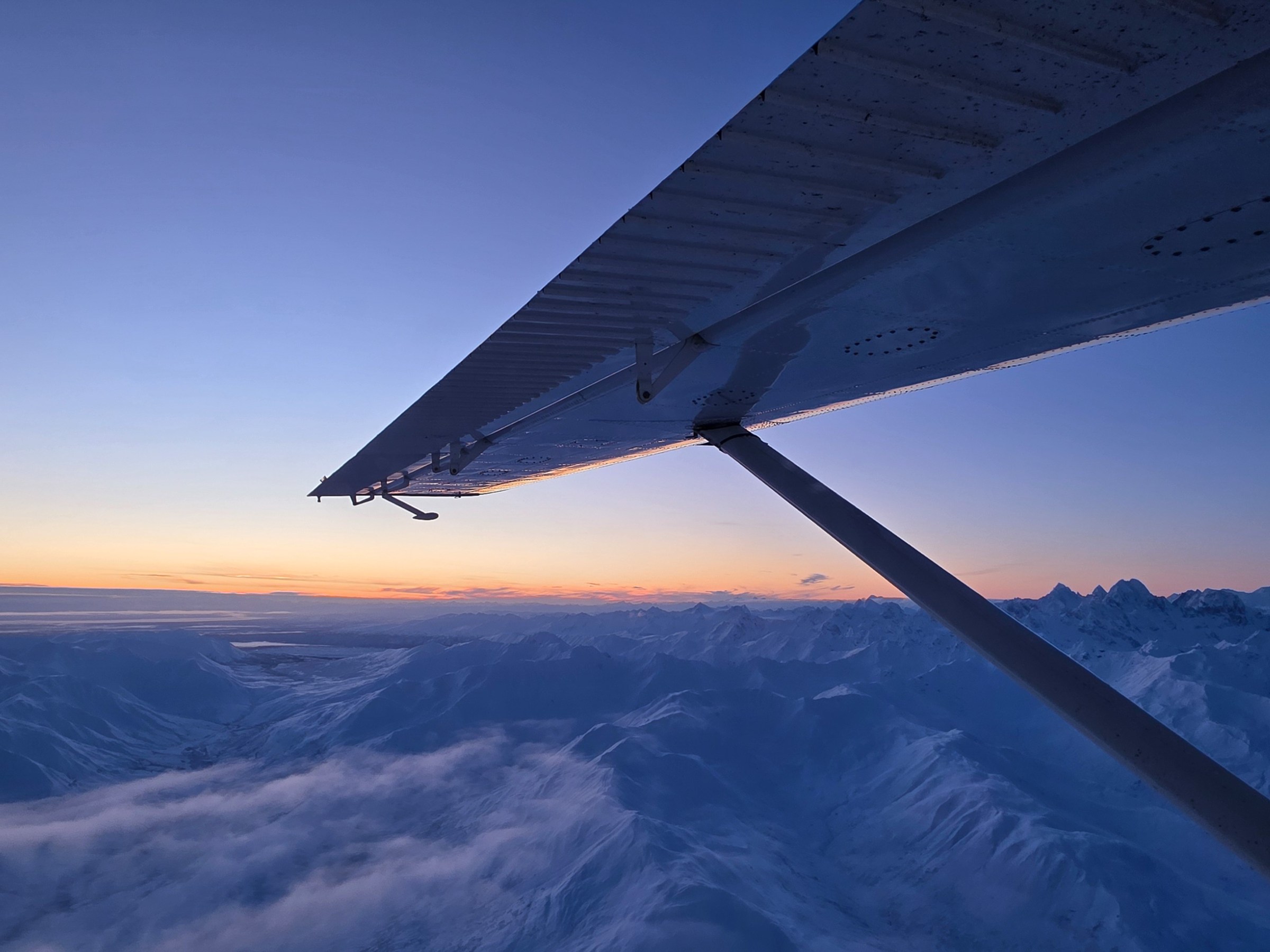 Airplane wing over snowy mountains at sunset with clear sky.
